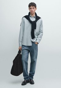 Camicia azzurra chiara con bottoni e jeans scuri, abbinati a un maglione nero appoggiato sulle spalle e a una borsa a spalla marrone scuro. Scarpe nere.