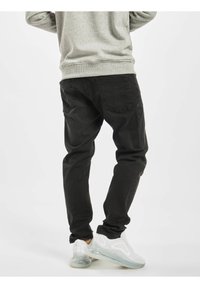 Osoba oblečená v černých džínách, šedé mikině a bílých teniskách s průhlednými podrážkami, stojící s překříženýma nohama a otočená zády.