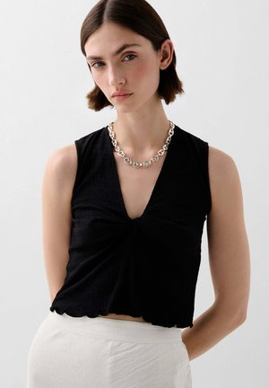 Top nero senza maniche con scollatura a V e design frontale annodato, caratterizzato da un tessuto testurizzato e orlo ondulato. Abbinato a una collana in argento.