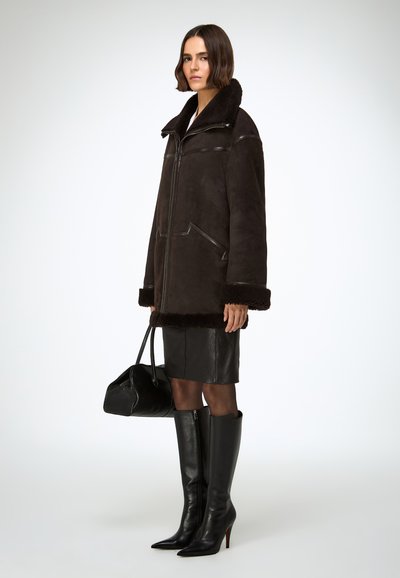 Veste en shearling marron foncé avec un col haut, fermeture éclair frontale et deux poches latérales. Associée à une jupe en cuir noire et des bottes noires montant jusqu'aux genoux.