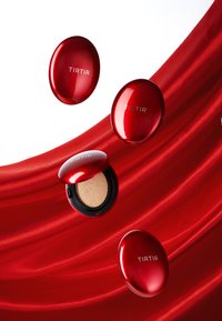 Red compact makeup case with TIRTIR branding. One open revealing a sponge, surrounded by additional closed compacts against a red satin backdrop.