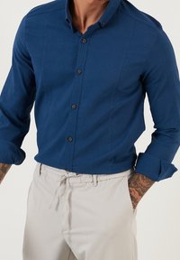 Niebieska koszula z guzikami, wykonana z teksturowanej tkaniny, z kołnierzykiem i długimi rękawami, zestawiona ze jasnymi spodniami.