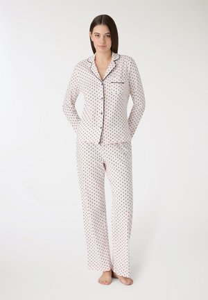 Donna con capelli castani lisci che indossa un pigiama a maniche lunghe rosa pallido con piccoli cuori neri, in piedi a piedi nudi su sfondo bianco.