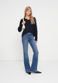 Pulôver de malha azul-marinho com um corte solto e decote em V, combinado com jeans claros de boca de sino. O conjunto é complementado por uma pequena bolsa branca e sapatos pretos de bico fino.