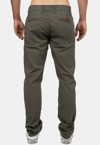 Homme portant un pantalon vert olive avec des détails de couture rouge, vu de dos, associé à des baskets Converse All Star blanches.