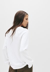 White long-sleeve shirt with a rounded neckline, soft texture, and loose fit. No visible patterns or hardware details. Model is facing sideways.