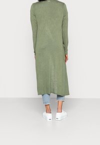 Largo vestido verde oliva con mangas largas y un dobladillo redondeado, combinado con pantalones vaqueros cortos de un azul claro y zapatillas de plataforma blancas.