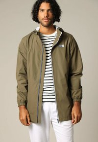 Olijfgroen waterdichte jas met ritssluiting, elastische manchetten en verstelbare capuchon; gedragen over een gestreept shirt en witte broek.