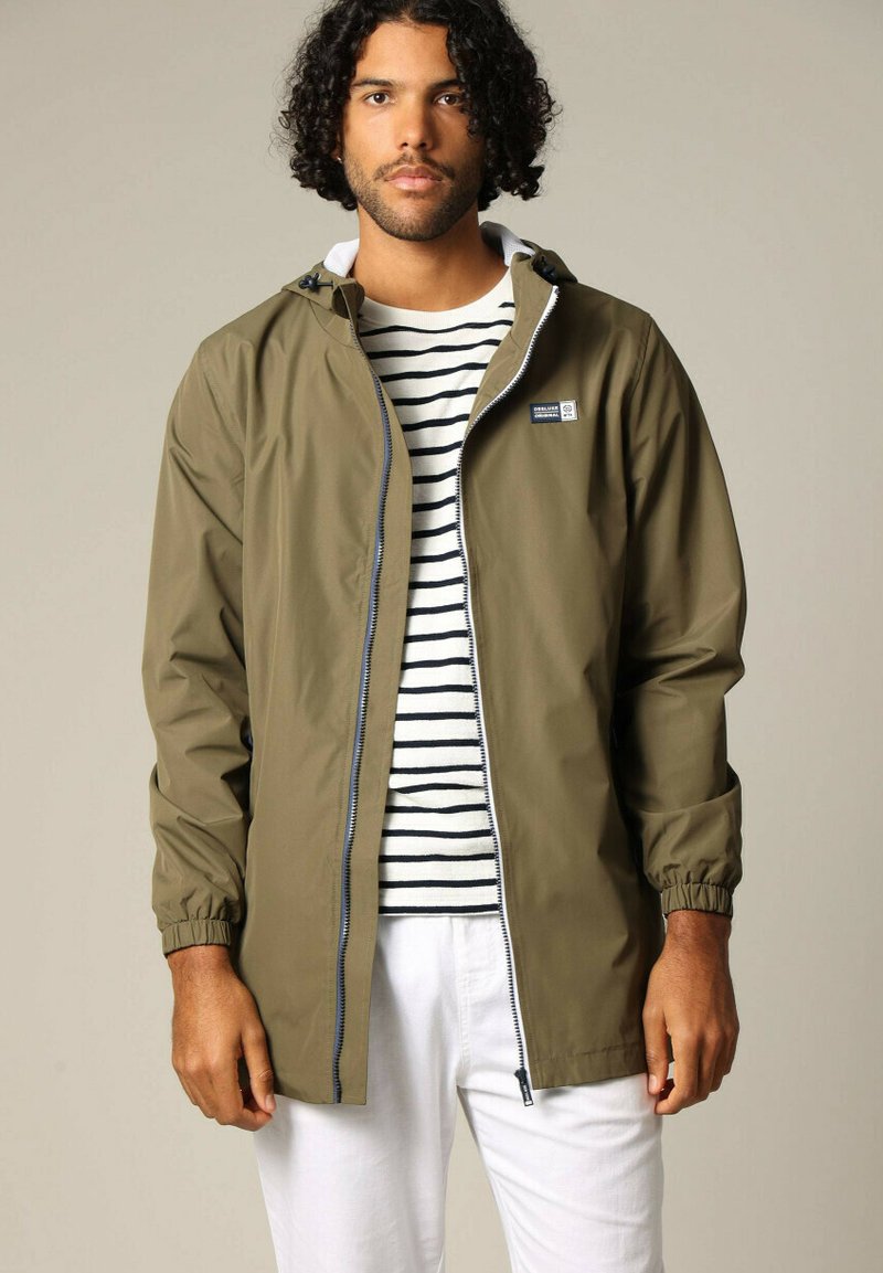 Olijfgroen waterdichte jas met ritssluiting, elastische manchetten en verstelbare capuchon; gedragen over een gestreept shirt en witte broek.