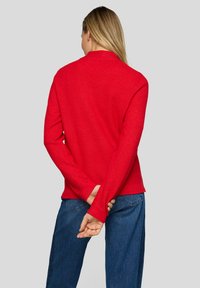 Frau mit langen blonden Haaren, die ein rotes langärmeliges Oberteil und blaue Jeans trägt, steht mit hinter dem Rücken verschränkten Händen und blickt weg.