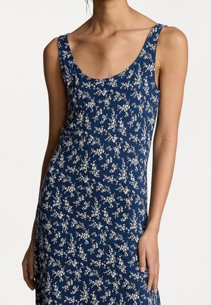 Kobieta ubrana w granatową sukienkę bez rękawów z białym kwiatowym wzorem, stojąca na tle jednolitego jasnego tła.