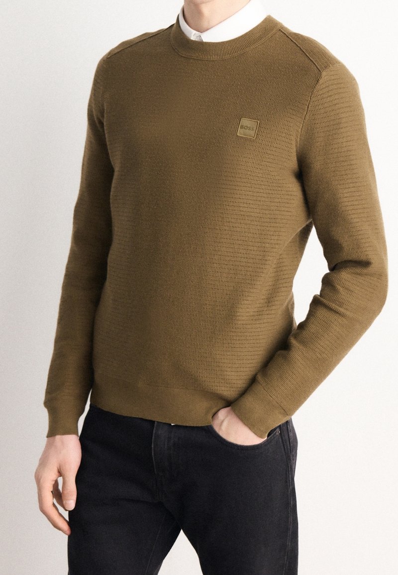 Homme portant un pull texturé vert olive par-dessus une chemise blanche à col, avec un jean noir, une main dans la poche, sur un fond uni.