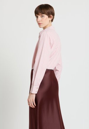 Donna con capelli corti castani che indossa una camicia a maniche lunghe rosa e bianca a righe infilata in una gonna di raso bordeaux scuro a vita alta, in piedi di profilo.