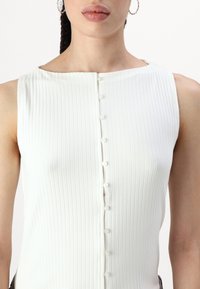Haut sans manches blanc avec une texture côtelée, présentant un décolleté arrondi et une patte de boutonnage à l'avant avec de petits boutons blancs.