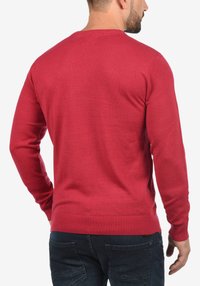 Pull-over rouge avec un col rond, des poignets et un ourlet côtelés, fabriqué en tissu tricoté texturé. Vue de dos montrant une silhouette ajustée.