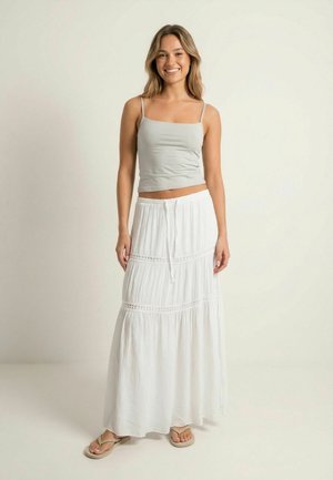 Usmievajúca sa žena v svetlosivej top s tenkými ramienkami, dlhej bielej vrstvenej sukni so šnúrkami a béžových žabkách stojaca pred jednoduchým pozadím.