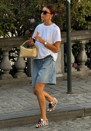 Vrouw met zonnebril, wit T-shirt, denimshorts en schoenen met koeienprint, die een ijsje en een beige handtas vasthoudt terwijl ze over een stenen pad loopt.