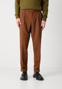 Braune gefütterte Hose mit taillierter Passform, aus glattem Stoff gefertigt; mit Knopfverschluss und umgeschlagenen Säumen, kombiniert mit schwarzen Slippern.