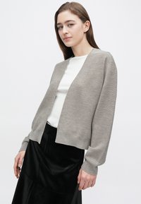 Cardigan grigio chiaro con vestibilità comoda, texture a coste, apertura frontale e maniche lunghe. Indossato sopra una maglia bianca e una gonna nera lucida.