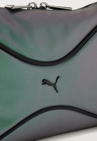 Αθλητική μαύρη τσάντα Puma με καμπυλωτές μαύρες ρίγες και διπλά ασημένια φερμουάρ πάνω σε μια λεία σκούρα γκρι επιφάνεια.