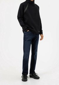 Sudadera negra de cuello alto con costuras laterales verdes, combinada con vaqueros azul oscuro y zapatillas negras. Ajuste casual, tela suave, diseño minimalista.