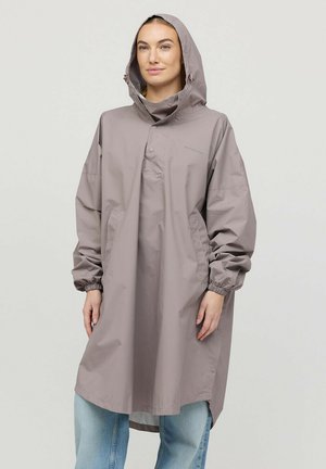 Vrouw draagt een oversized lichtgrijze regenjas met capuchon over lichtblauwe spijkerbroek, staand tegen een effen achtergrond, handen ontspannen.