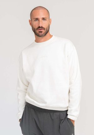 Man with short hair and beard wearing white sweatshirt and dark gray pants, posing with hands in pockets against plain background.
