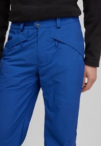 Gros plan d'une personne portant un pantalon bleu avec des poches avant inclinées et un haut noir à manches longues, main détendue le long du corps.