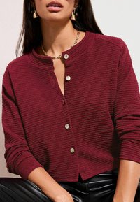 Cardigan burgund texturat cu guler rotund, prevăzut cu nasturi aurii și mâneci lungi, asortat cu pantaloni negri eleganți.