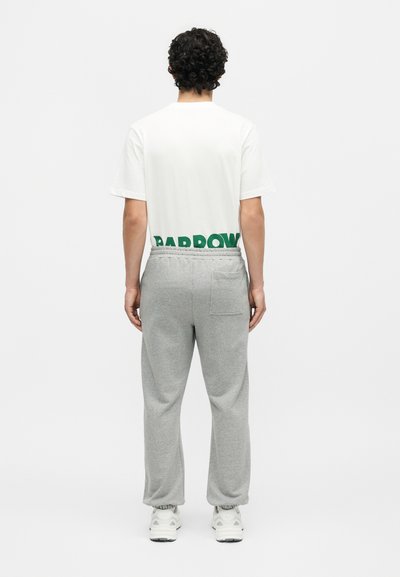 Άνδρας με σγουρά σκούρα μαλλιά στέκεται γυρισμένος προς τα πίσω φορώντας λευκό μπλουζάκι με πράσινο κείμενο στο κάτω μέρος και γκρι φόρμα με τσέπη.