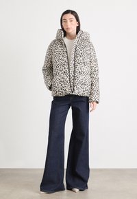 Eine Frau steht frontal, trägt eine Daunenjacke mit Leopardenmuster, ein helles Oberteil, weit ausgestellte Jeans und helle Schuhe, vor einem neutralen Hintergrund.