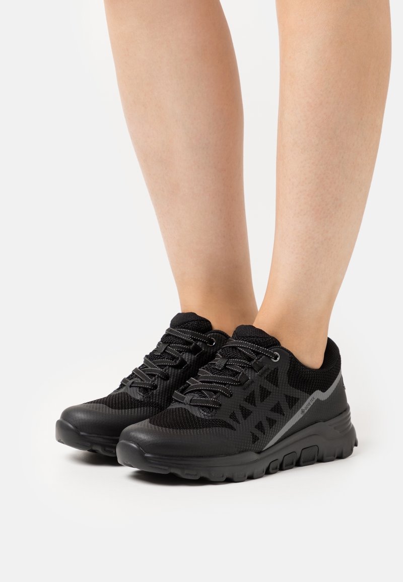 Jambes portant des baskets de sport noires avec tige en mesh et synthétique, côtés à motifs et semelles épaisses et rembourrées sur fond blanc.