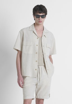 Beige shirt met korte mouwen, knoopsluiting en twee voorkruiden, gecombineerd met bijpassende shorts. gedragen met donkere rechthoekige zonnebril.