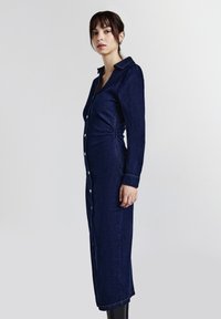 Robe chemise en denim, bleu foncé, à manches longues, fermeture à boutons, détail froncé sur le côté, coupe ajustée, longueur cheville, design minimaliste.