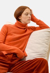 Turtleneck-Pullover aus leuchtend orangefarbenem Strick mit gerippten Bündchen. Kombiniert mit strukturierten, rostfarbenen Cordhosen vor einem weißen Kissenhintergrund.