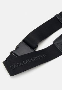 Črn nastavljiv trak iz trpežnega najlona, z mat črno plastično sponko in vtisnjenim blagovno znamko 'KARL LAGERFELD'.