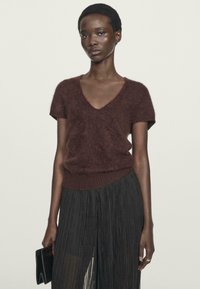 Maglione corto a maniche corte marrone e peloso con scollo a V, abbinato a una lunga gonna nera plissettata. La modella tiene una pochette nera.