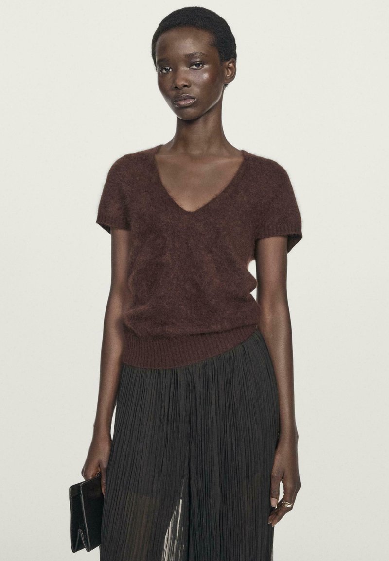 Maglione corto a maniche corte marrone e peloso con scollo a V, abbinato a una lunga gonna nera plissettata. La modella tiene una pochette nera.