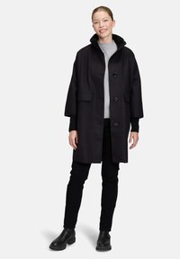 Czarny wełniany płaszcz z wysokim kołnierzem, guzikami z przodu i dużymi kieszeniami. Nosić z szarym swetrem i czarnymi spodniami, zestawiony z czarnymi butami.