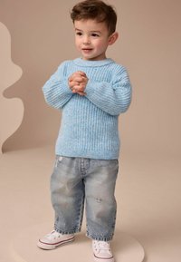 Jasnoniebieski sweter z dzianiny o textured wzorze, zestawiony z przetartymi jasnymi dżinsami i białymi trampkami z czerwonymi akcentami.