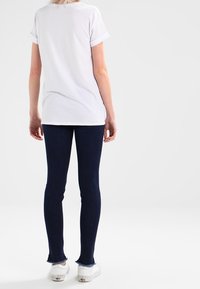 Weißes T-Shirt mit aufgerollten Ärmeln, kombiniert mit dunkelblauen Skinny-Jeans mit Fransen-Saum, ergänzt durch weiße Sneaker.