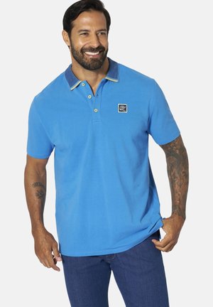 Smiling man with beard wearing bright blue polo shirt with contrasting collar and dark blue jeans, showing tattooed arms, holding shirt hem.