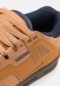 Brown leather sneaker with textured accents, blue inner lining, and thick beige laces. Features perforated areas and a logo tag on the side.