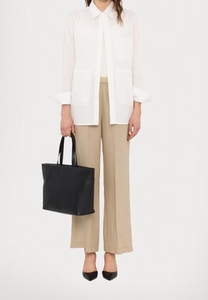 Personne portant une chemise blanche boutonnée, un pantalon beige à jambes larges, des chaussures noires pointues, tenant un sac fourre-tout en cuir noir avec des poignées.