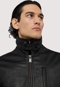 Svart läderjacka med hög krage, dragkedja framtill och tryckknappdetaljer. Strukturerad yta och sidofickor syns.