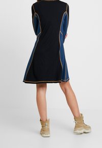 Knälång klänning med blå och svarta paneler, accentuerad med guldstygn, i kombination med beige ankelskor med en strukturerad yta.