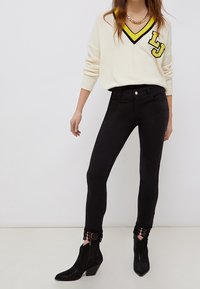 Žena oblečená v krémovém svetr s V výstřihem a žluto-černým lemem, černých skinny džínách s krajkovými manžetami a černých kotníkových botách na podpatku.