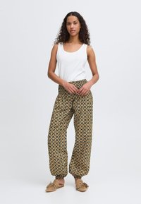 Weißes ärmelloses Oberteil, gemusterte gelbe und schwarze locker sitzende Hose mit elastischen Bündchen, ergänzt durch tanfarbene Fransen-Schlappen.