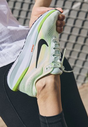 Personne étirant la jambe portant une chaussure de course Nike vert clair et blanche avec un swoosh noir et le texte "Air" sur la semelle.