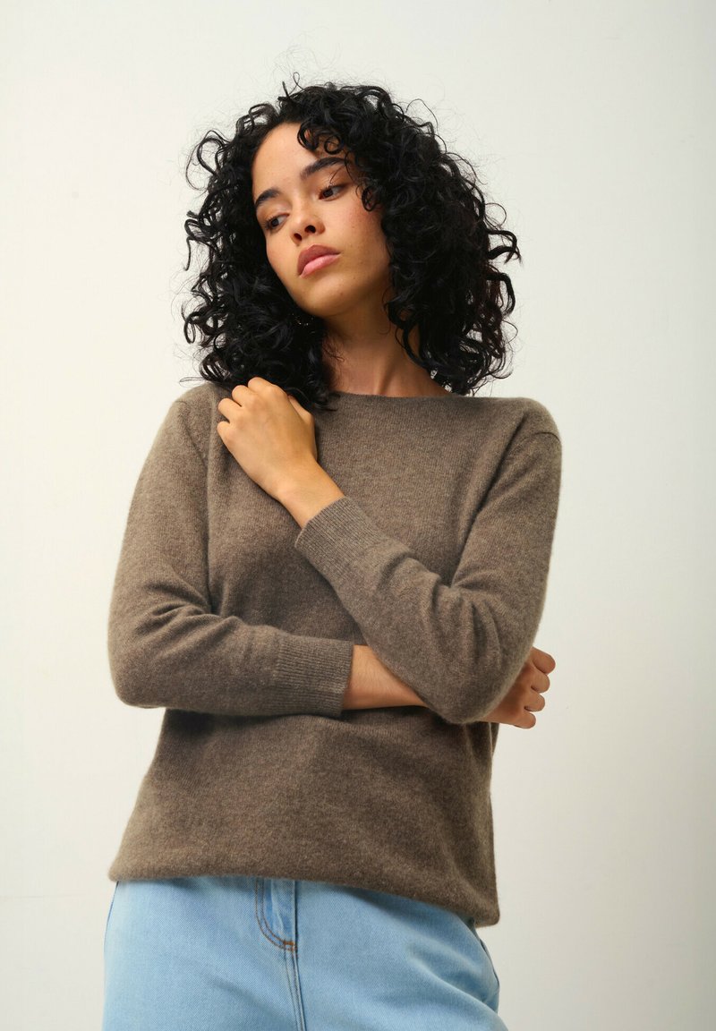 Kvinna med lockigt svart hår, iförd en brun tröja och ljusblå jeans, står framför en enkel ljus bakgrund.